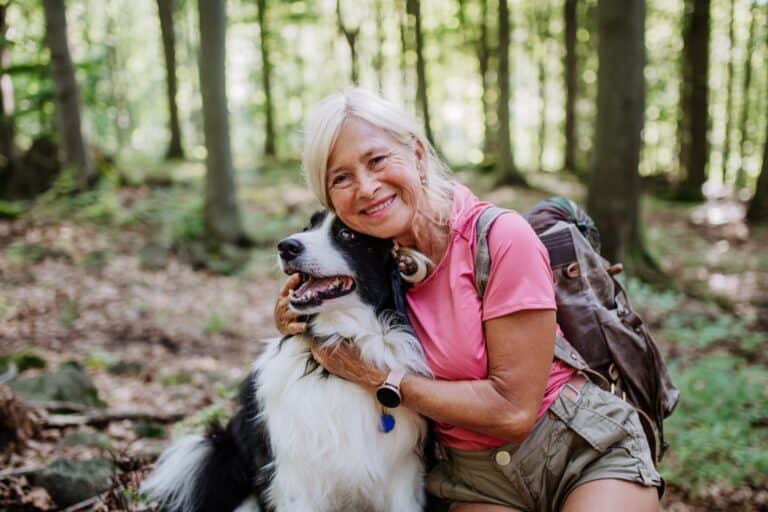 A woman walking in the woods with her border collie, petting it and leaning against it.